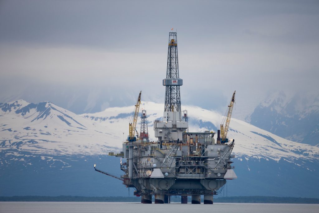 An offshore oil drilling platform stands in calm water with snow-covered mountains and a cloudy sky behind it. Several cranes are visible on the rig in this high-quality image, perfect for Adobe Stock or other royalty-free stock images collections.