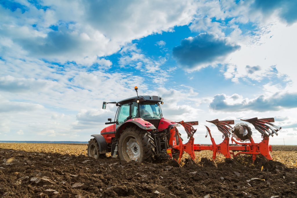 A red tractor pulls a plow through a freshly tilled field under a partly cloudy blue sky, preparing the soil for planting.