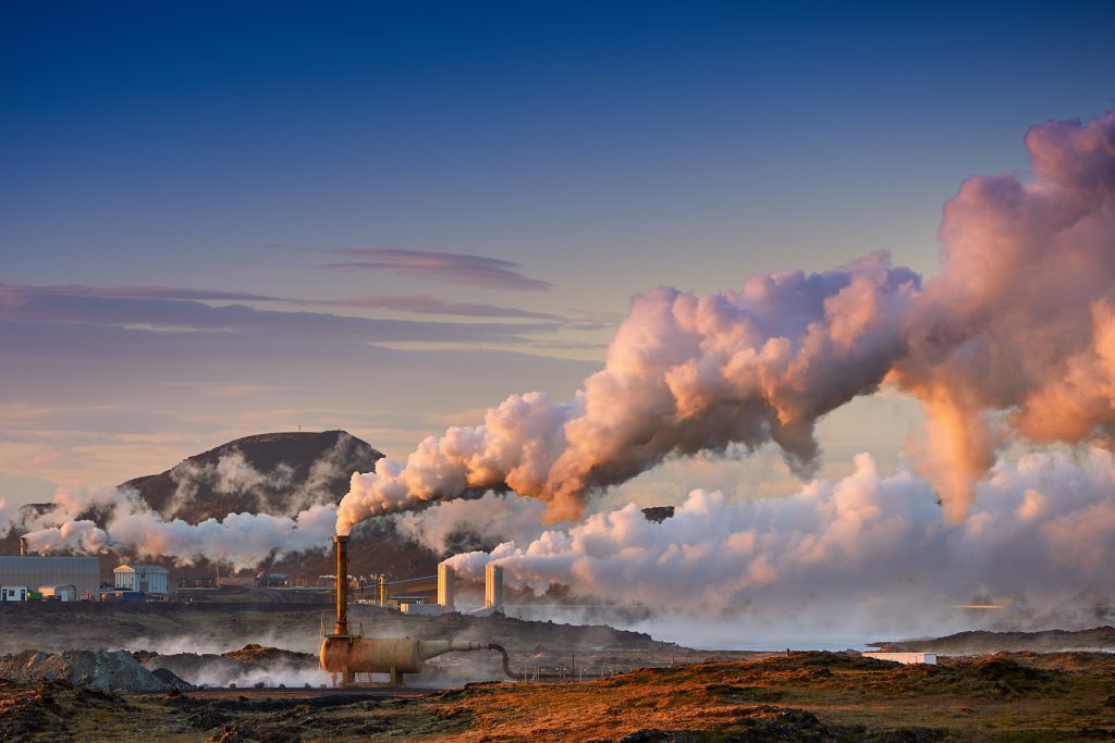Industrial facility emits thick white steam or smoke into the sky at sunset, with a mountain in the background and colorful clouds, with a geothermal energy plant in a rugged landscape.