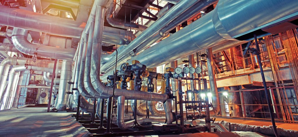 Large industrial facility interior with numerous metal pipes, gauges, and valves. The scene is well-lit, and the structures have a metallic sheen, suggesting a factory or power plant environment.
