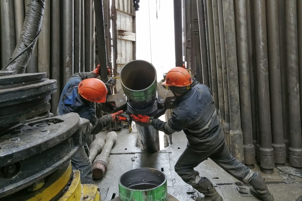 Two drilling crew workers in orange helmets and safety gear dismantle the bottom of the drill string by unscrewing the spiral calibrator and working with a machine key on the rotary platform of the drilling rig.