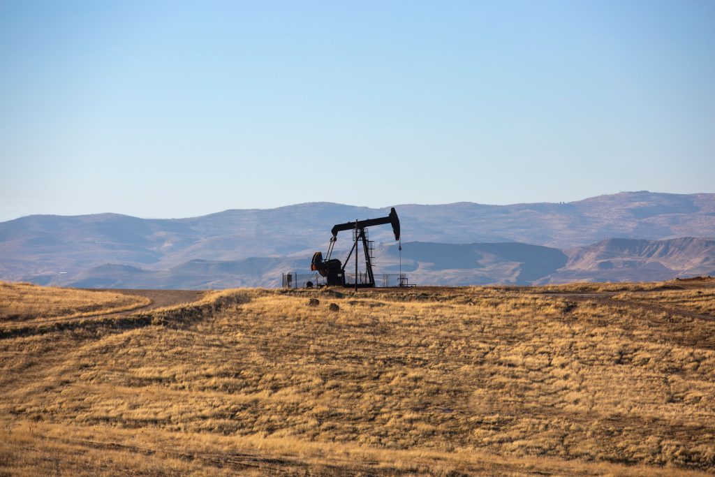 An oil pumpjack operates in a dry, grassy field with distant rolling hills and a clear blue sky in the background.