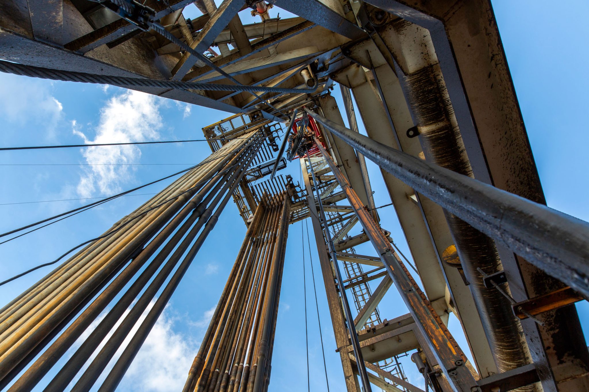 Upward view from the base of an oil drilling rig with metal pipes and structures extending into the sky, framed against blue sky and clouds.